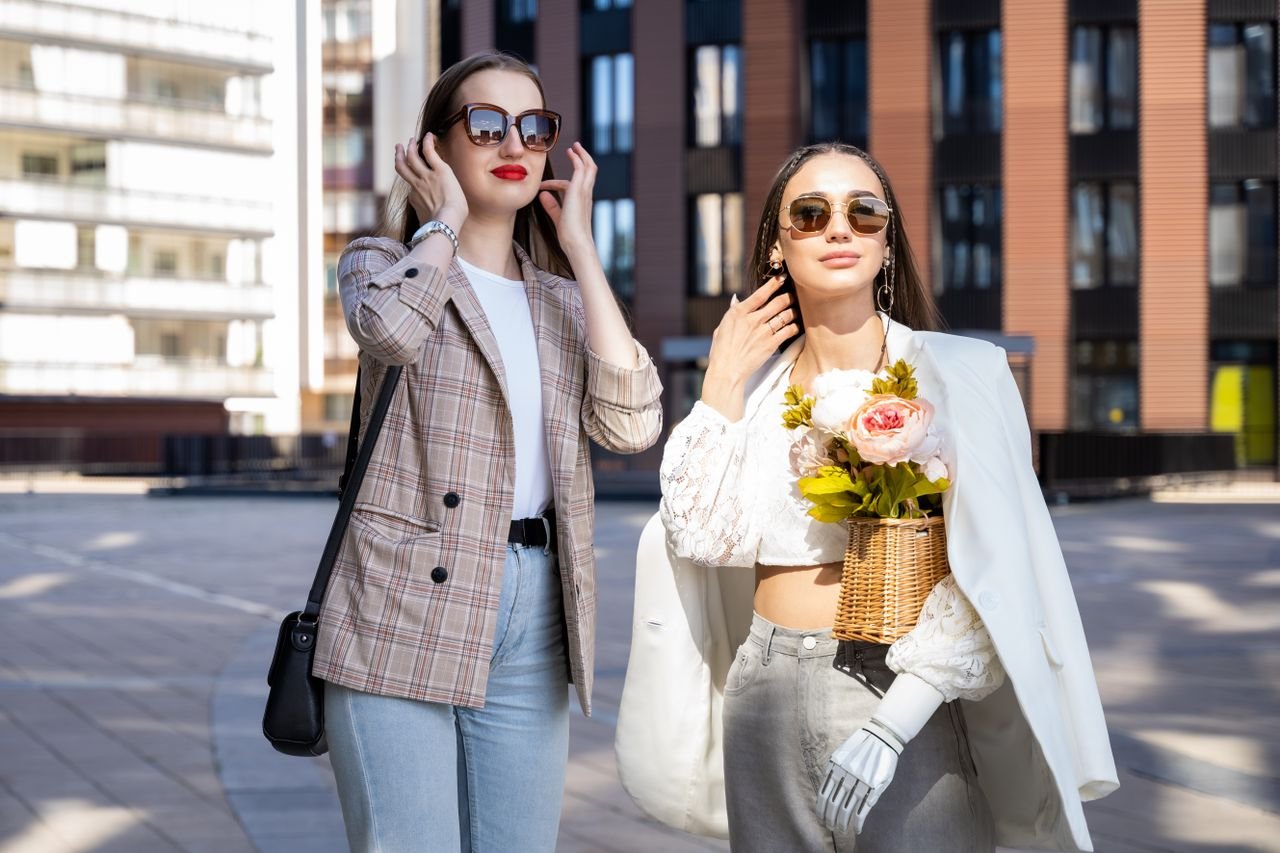 Two stylish women enjoy a sunny day in the city wearing chic outfits and sunglasses.